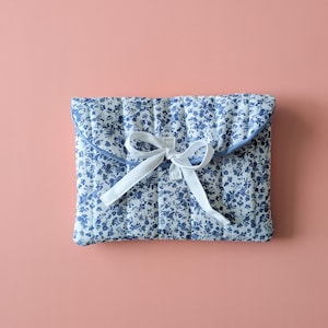 以下が含まれることがあります： 白地に青い花柄のキルティングクラッチ。白いリボンで結ばれています。縁は無地の青い生地でトリミングされています。背景は無地のピンク色です。