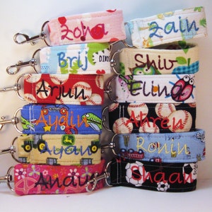 May include: A stack of colorful fabric keychains with personalized names, including "Zora", "Brij", "Arjun", "Aydin", "Aurelio", "Anav", "Zain", "Shiv", "Elina", "Ahren", "Ronin", and "Shaan".
