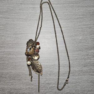 May include: A long, antique gold-toned necklace with a pendant featuring a cluster of decorative elements. The pendant includes a large, faceted amber-colored gem, faux pearls, and detailed leaf charms. The chain is a delicate link style.