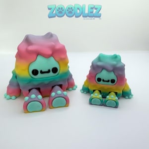 May include: Two colorful monster figurines with rainbow-colored bodies and teal faces, feet, and hands. The larger figure is on the left, and the smaller one is on the right. The text "ZOOODLEZ" is at the top.