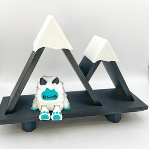 May include: A whimsical, black and white mountain-shaped shelf with two peaks, each topped with a white triangle. A cute, white and blue yeti figurine sits below the peaks. The shelf is designed to hold small items.