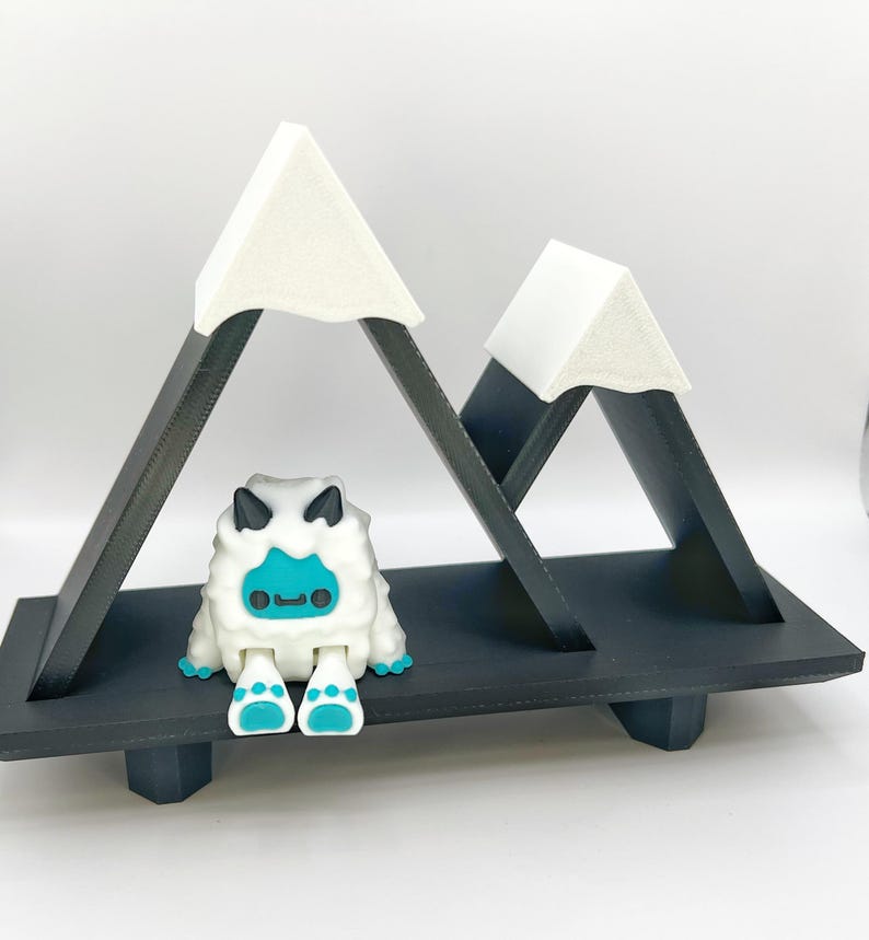 May include: A whimsical, black and white mountain-shaped shelf with two peaks, each topped with a white triangle. A cute, white and blue yeti figurine sits below the peaks. The shelf is designed to hold small items.