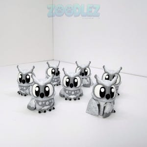 May include: A collection of six small, gray and white speckled owl figurines with large black eyes and stylized features. Each owl has a textured surface and sits in a seated position. The word ZOODLEZ is printed in blue at the top center.