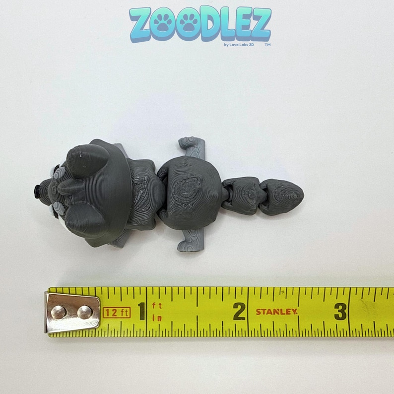 May include: A gray 3D-printed articulated raccoon toy, approximately 3 inches long, with a white face and a segmented tail. The toy is measured with a yellow tape measure. The Zoodlez logo is visible in the upper left corner.