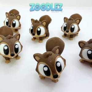 May include: Several 3D-printed cartoon squirrel figurines in shades of brown and tan are arranged on a white surface. The squirrels have large, expressive eyes and fluffy tails. The Zoodlez logo is visible in the top center.