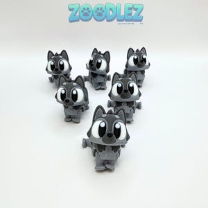 May include: A collection of gray wolf figurines with large, expressive eyes. The figurines are arranged in a group, with the text "Zoodlez" in blue above them. The wolf figures have a cartoonish design.