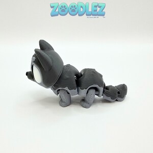 May include: A gray articulated cat toy with white accents, featuring a detailed design. The toy is in a playful pose, with the text "Zoodlez" in blue above. The toy is made by Love Labs 3D.