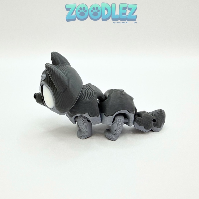 May include: A gray articulated cat toy with white accents, featuring a detailed design. The toy is in a playful pose, with the text "Zoodlez" in blue above. The toy is made by Love Labs 3D.