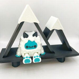 May include: A whimsical, white, furry creature with a turquoise face and feet sits on a black shelf beneath two triangular mountain peaks. The mountains have white tops and black supports. The creature has black horns and a small, smiling mouth.