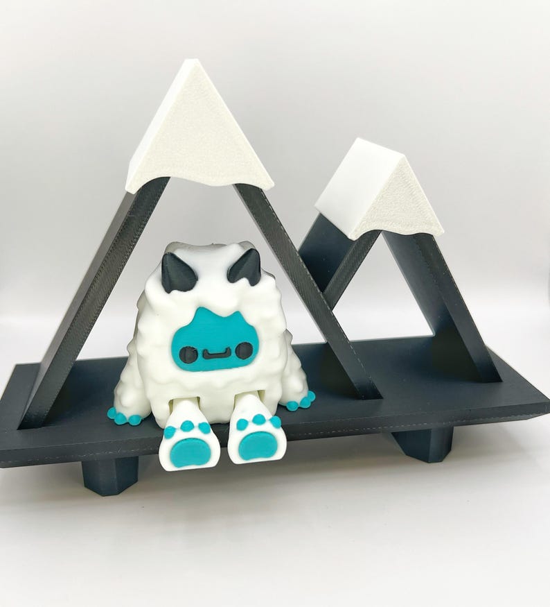 May include: A whimsical, white, furry creature with a turquoise face and feet sits on a black shelf beneath two triangular mountain peaks. The mountains have white tops and black supports. The creature has black horns and a small, smiling mouth.