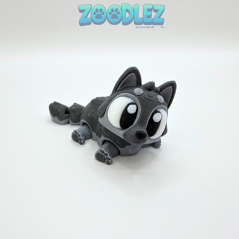 May include: A 3D-printed, gray and white raccoon figurine with large, expressive eyes. The figurine is lying down, with its paws and tail visible. The text "Zoodlez" is in the upper left corner.