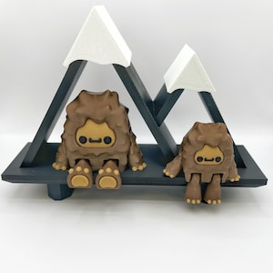 May include: A whimsical display featuring two brown Bigfoot figurines with tan accents, seated on a dark gray platform. The backdrop includes two white mountain peaks, creating a playful, nature-inspired scene. The figurines have a cartoonish design.