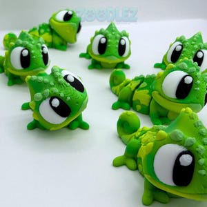 May include: A collection of small, articulated green chameleon figurines with large white eyes and black pupils. Each lizard features a textured, bumpy surface and a bright lime and forest green color scheme. The word ZOO DLEZ is visible in the background.