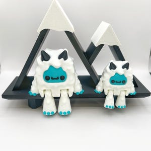 May include: Two white and teal yeti figurines with black horns and teal faces, standing on a black shelf in front of a black mountain-shaped structure with white snowy peaks. The figurines have teal feet and hands.
