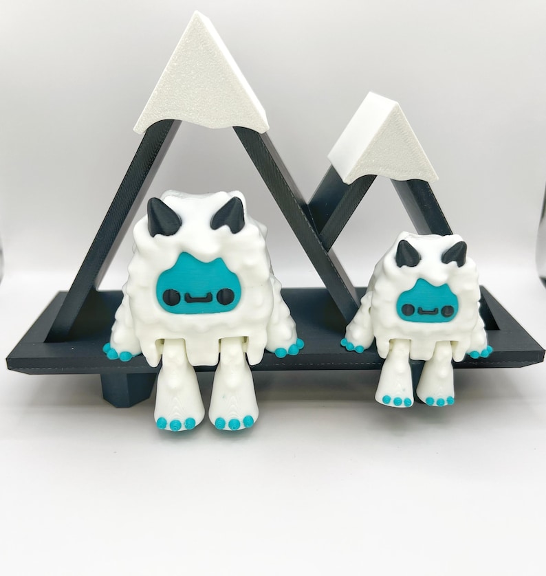May include: Two white and teal yeti figurines with black horns and teal faces, standing on a black shelf in front of a black mountain-shaped structure with white snowy peaks. The figurines have teal feet and hands.