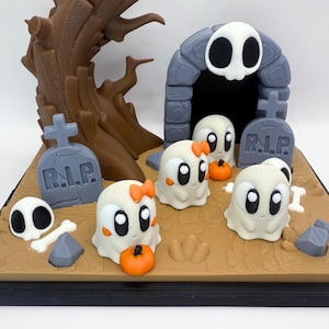 May include: A Halloween-themed diorama featuring cartoon ghosts, pumpkins, tombstones, and a skull-shaped archway. The ghosts have large eyes and orange bows. The scene is set on a brown base with a tree and bone accents.