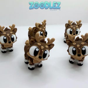 May include: Several brown and tan deer figurines with large, expressive eyes and antlers. The figurines have white accents on their feet and around their eyes. The word "Zoodlez" is written in blue and white bubble letters at the top of the image.
