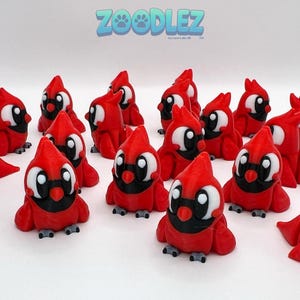 May include: A collection of red cardinal figurines with black and white facial details. The figurines have a 3D-printed appearance and are arranged on a white surface. The logo "Zoodlez" is visible in the background.