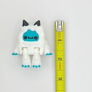 May include: A white, articulated toy figure resembling a yeti, with a blue face and feet, and black ears. The figure is approximately 2 inches tall, as measured by a yellow tape measure.