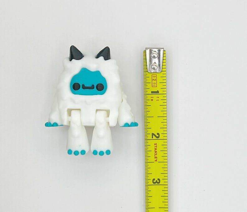 May include: A white, articulated toy figure resembling a yeti, with a blue face and feet, and black ears. The figure is approximately 2 inches tall, as measured by a yellow tape measure.