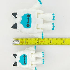 May include: Two white Yeti-like figurines with black horns and teal faces and feet. The figurines are approximately 2 inches long. A yellow Stanley measuring tape is placed next to the figurines.