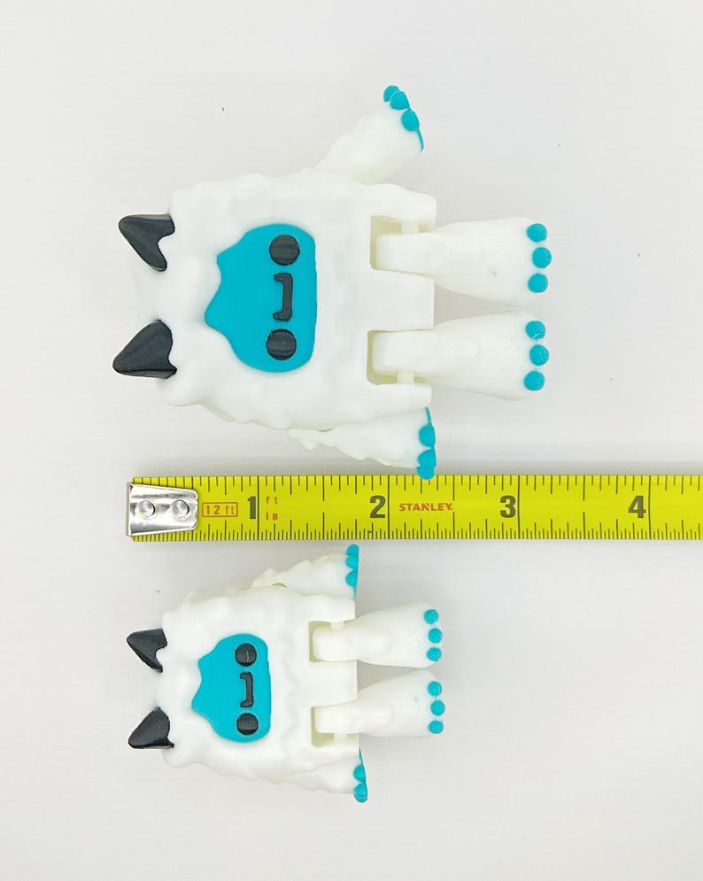 May include: Two white Yeti-like figurines with black horns and teal faces and feet. The figurines are approximately 2 inches long. A yellow Stanley measuring tape is placed next to the figurines.