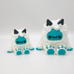 May include: Two white, fluffy monster figurines with teal faces and feet, and black horns. The larger figure has two feet, while the smaller has two feet. The figures have a cartoonish design.