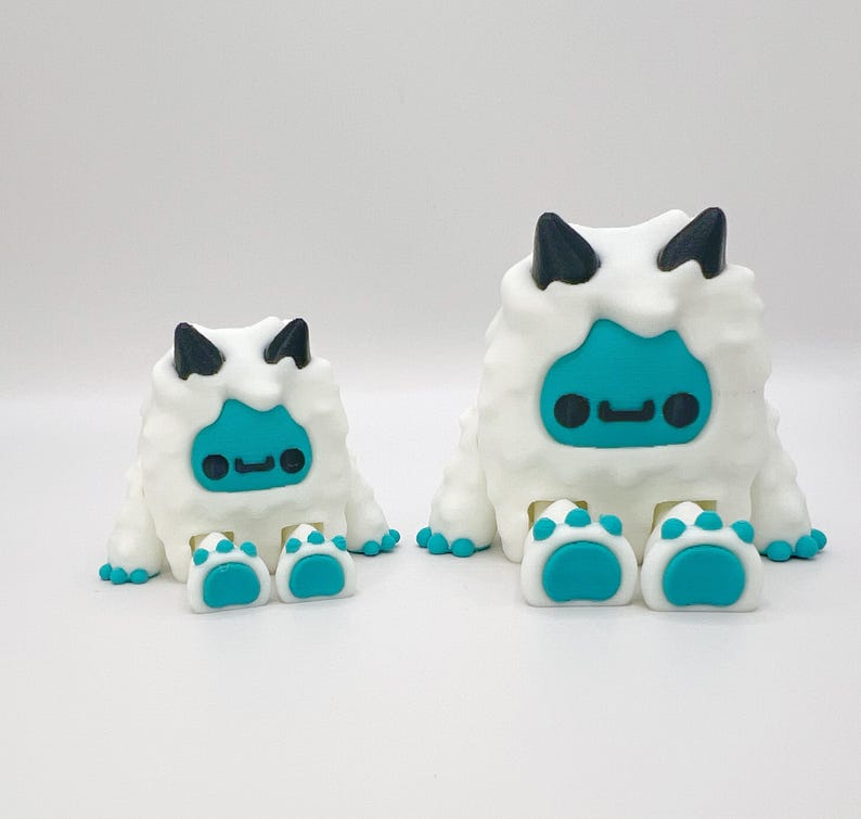 May include: Two white, fluffy monster figurines with teal faces and feet, and black horns. The larger figure has two feet, while the smaller has two feet. The figures have a cartoonish design.