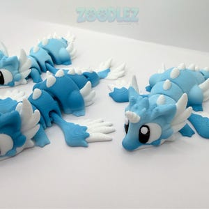 May include: Three articulated dragon figurines in shades of light and dark blue with white accents, including horns, wings, and fins. Each creature features large black and white eyes and a segmented body design. The text ZOODLEZ appears at the top.