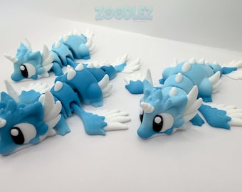 May include: Three articulated dragon figurines in shades of light and dark blue with white accents, including horns, wings, and fins. Each creature features large black and white eyes and a segmented body design. The text ZOODLEZ appears at the top.