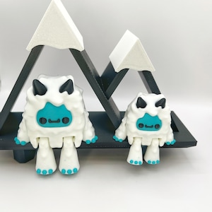 May include: Two white yeti figurines with turquoise faces and feet, and black horns. They are positioned in front of a black wooden mountain-shaped shelf with white mountain peaks.