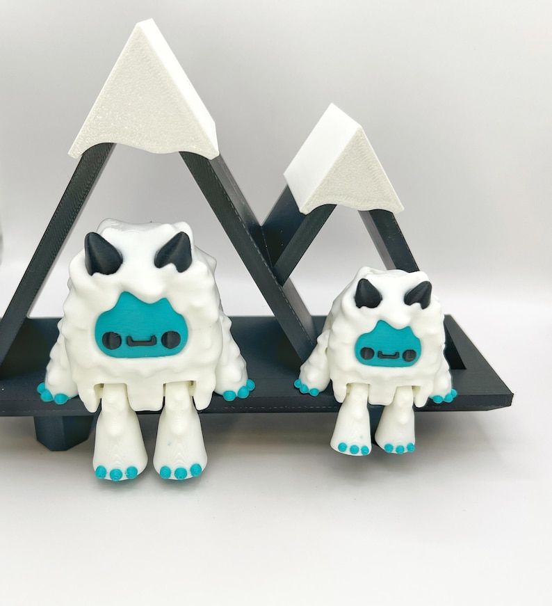 May include: Two white yeti figurines with turquoise faces and feet, and black horns. They are positioned in front of a black wooden mountain-shaped shelf with white mountain peaks.