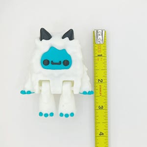 May include: A white, articulated toy figure resembling a yeti, with black horns and blue facial features. The figure has blue accents on its feet and hands. A yellow measuring tape is positioned next to the toy, showing a height of approximately 3 inches.