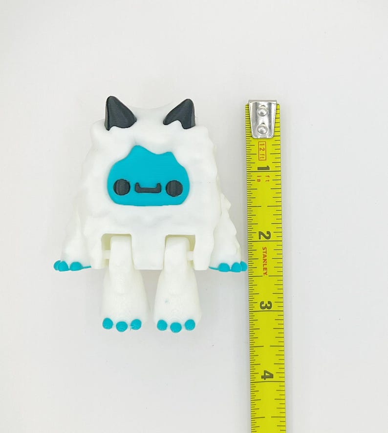 May include: A white, articulated toy figure resembling a yeti, with black horns and blue facial features. The figure has blue accents on its feet and hands. A yellow measuring tape is positioned next to the toy, showing a height of approximately 3 inches.