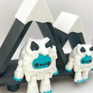 May include: Two white yeti figurines with black horns and teal faces and feet stand in front of a black and white mountain range. The yeti have a friendly expression. The mountain range is made of black triangles with white peaks.