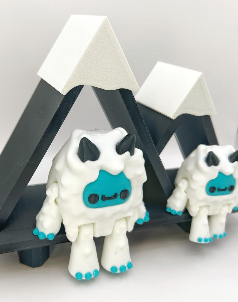 May include: Two white yeti figurines with black horns and teal faces and feet stand in front of a black and white mountain range. The yeti have a friendly expression. The mountain range is made of black triangles with white peaks.