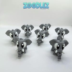 May include: A collection of gray 3D-printed elephant figurines with large white eyes and tusks. The figurines are arranged on a white surface. The text "Zoodlez" is displayed in blue above the elephants.
