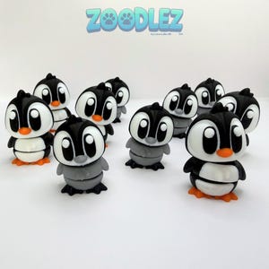 May include: A collection of penguin figurines in black, white, gray, and orange. The penguins have large white eyes, orange feet, and are arranged on a white surface. The word "ZOOZLEZ" is displayed in blue above the penguins.