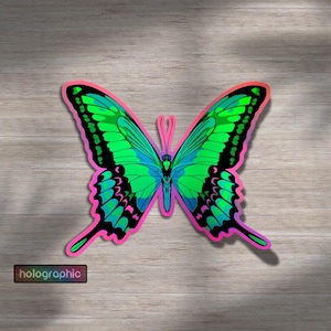 Puede incluir: Una pegatina holográfica de mariposa con un diseño vibrante. La mariposa presenta colores verdes, azules y rosas. La palabra "holographic" está impresa en una pequeña etiqueta rectangular. La pegatina está sobre una superficie de madera.