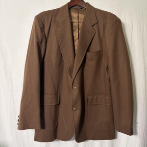May include: A brown blazer with two front pockets and gold buttons. The blazer is hanging on a wooden hanger against a white background. The blazer has a notched lapel and a single-breasted closure.