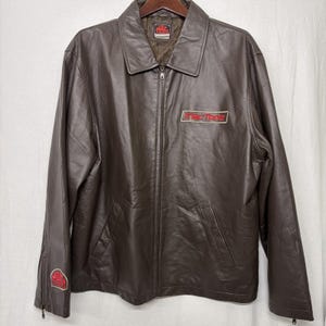 May include: Brown leather jacket with a full zipper closure, a collar, and two front pockets. The jacket features the "Mac Tools" logo in red and white on the chest and sleeve. The jacket is hanging on a wooden hanger.