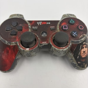 May include: A wireless game controller featuring a gray digital camouflage pattern with red smoke accents. The center displays the WWE 2K14 logo and Skinit branding. The right side features a portrait of a wrestler, while standard buttons are present.