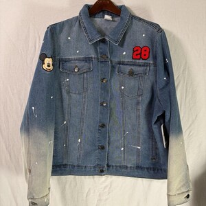 May include: A blue denim jacket with a button-up front, featuring a Mickey Mouse patch on the left shoulder and the number "28" on the right. The jacket has white paint splatters and a faded ombre effect on the sleeves.