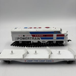 May include: A white and blue Powertrain model train with red accents and the text "The All American Powertrain." Below is a white Loop-the-Loop Cars flatbed car. The train is on a black track.
