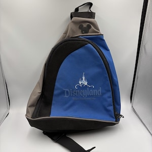 May include: A blue and gray sling backpack with the Disneyland logo. The bag features a black mesh panel, a black strap, and a Mickey Mouse silhouette. The bag is designed to be worn across the body.