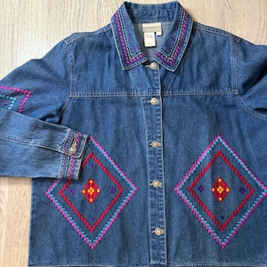 May include: A denim jacket with colorful embroidered details. The jacket features a collar and cuffs with vibrant stitching in shades of purple, pink, and teal. The front of the jacket showcases two large diamond-shaped embroidered patterns in red, pink, and teal.