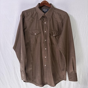 May include: A brown long-sleeve shirt with a button-down front, two chest pockets, and a pointed collar. The shirt has white buttons and a Western-style design. The shirt is hanging on a wooden hanger.