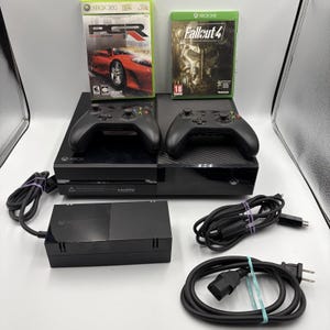 May include: A black Xbox console with two controllers, power supply, and game discs. The game discs are for PGR and Fallout 4. The controllers are black and the console is on a white surface.