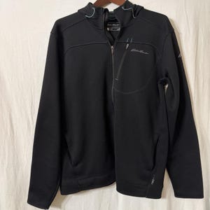 May include: Black zip-up hooded jacket with the Eddie Bauer logo on the chest. The jacket has a front zipper, two side pockets, and a small chest pocket. The jacket has a hood and a small blue accent on the hood.