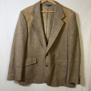May include: A brown tweed blazer with tan suede shoulder patches and elbow patches. The blazer has two front pockets, a two-button closure, and three buttons on each cuff. The jacket is hanging on a wooden hanger.